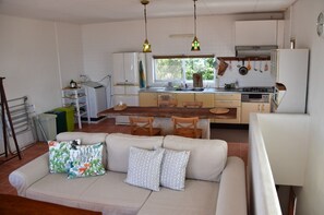 A compact kitchen with a dining area, featuring a sofa with patterned cushions, a wooden table, and a window providing natural light.