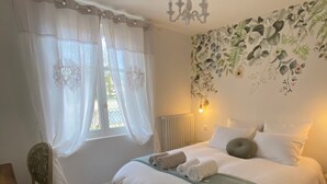 Une chambre à coucher avec un lit, des rideaux blancs, une chaise et un mur orné d’un motif floral.