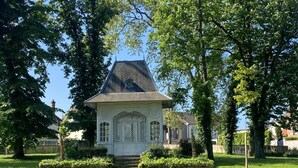Un petit bâtiment blanc, de plain-pied, avec un toit à forte pente, entouré d’une pelouse verdoyante et d’arbres.