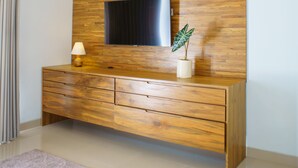 A modern living room with a wooden wall panel, a flat-screen TV mounted above a wooden cabinet, a lamp, and a potted plant. - Rapuan Cili Villa by Ilys Collection (Ubud)