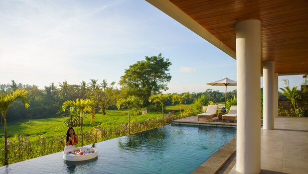 A pool area with a woman sitting on a floating platform, surrounded by lush greenery and a clear sky. - Rapuan Cili Villa by Ilys Collection (Ubud)