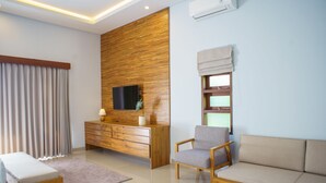 A modern living room with a wooden accent wall, a flat-screen TV, a sofa, a chair, and a small bedside table with a lamp. - Rapuan Cili Villa by Ilys Collection (Ubud)