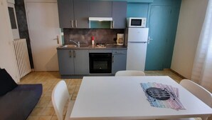 A compact kitchen with a sink, stove, microwave, and refrigerator. There is a dining table with chairs and a black couch in the adjacent living area.