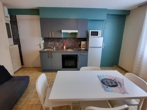 A compact kitchen with a sink, stove, microwave, and refrigerator. There is a dining table with chairs and a black couch in the adjacent living area.