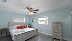 Une chambre à coucher comprenant un lit, un ventilateur de plafond, une table de chevet, une commode et deux fenêtres.