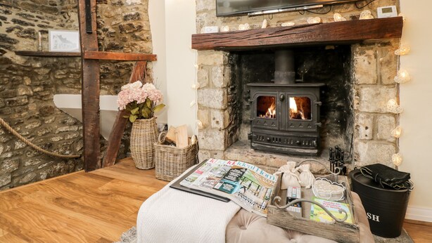 A cozy room with a stone wall, a fireplace, a television, and a wooden table with magazines and a basket. - Corner Cottage (Malmesbury)
