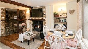 A cozy room with a stone fireplace, a television mounted above it, and a dining table set for four. - Corner Cottage (Malmesbury)