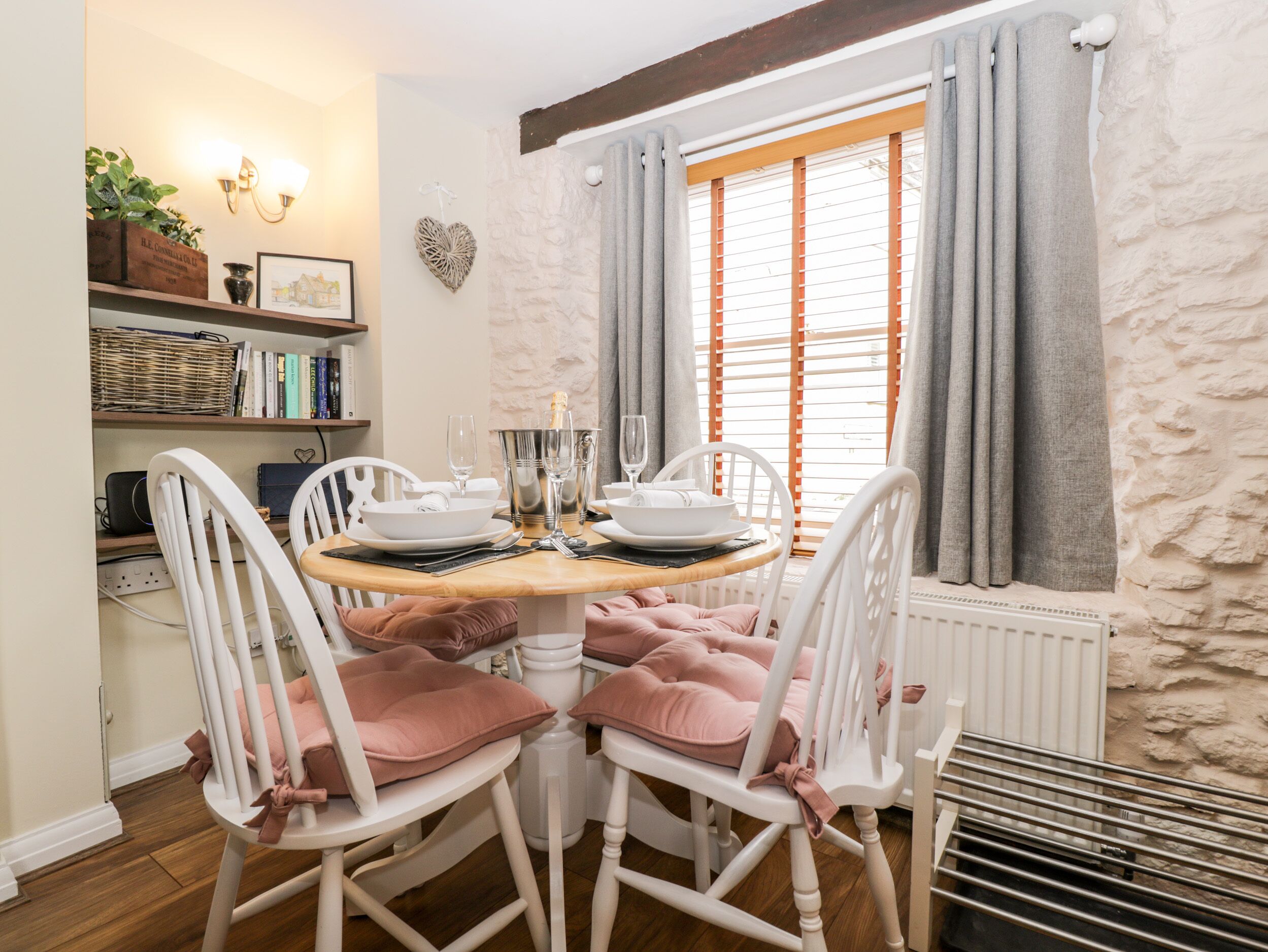 A dining area with a round table set for four, white chairs with pink cushions, a white wall with a shelf holding books and decorative items, and a window with grey curtains.