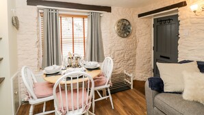 A dining area with a wooden table set for four, white chairs, a clock on the wall, and a stone wall. - Corner Cottage (Malmesbury)
