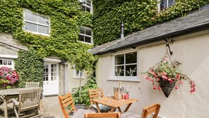 A patio with a wooden table and chairs, a hanging flower basket, and a stone wall with ivy. - Corner Cottage (Malmesbury)