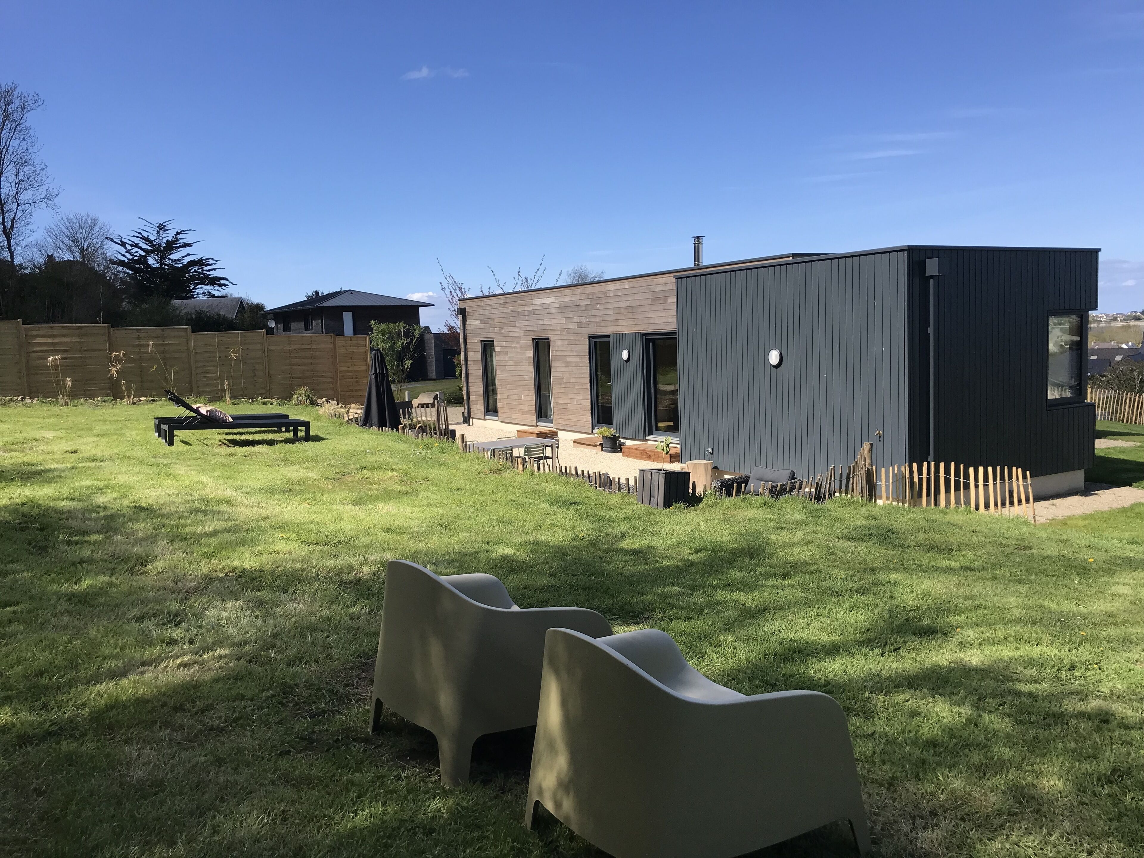 Een modern buitengebied met een houten terras, een lounge stoel en een klein gebouw met een donkere gevel.