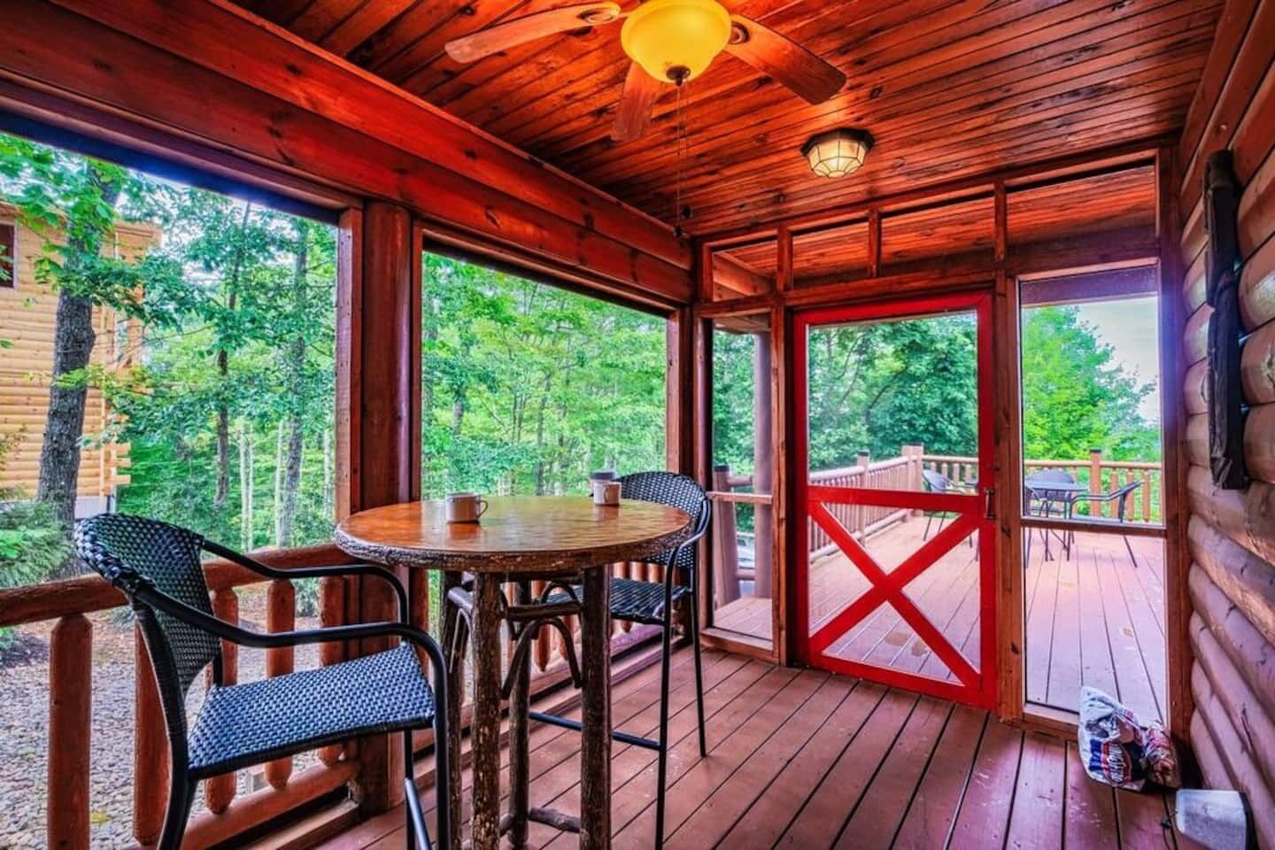 A wooden deck with a round table and chairs, large windows, and a view of the surrounding forest.