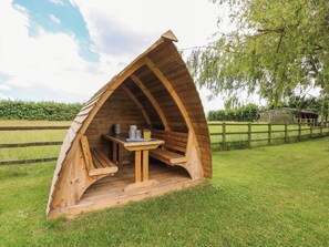 A wooden tent-like structure with a table and chairs inside, surrounded by a grassy area and a wooden fence. - The Granary (Boston)