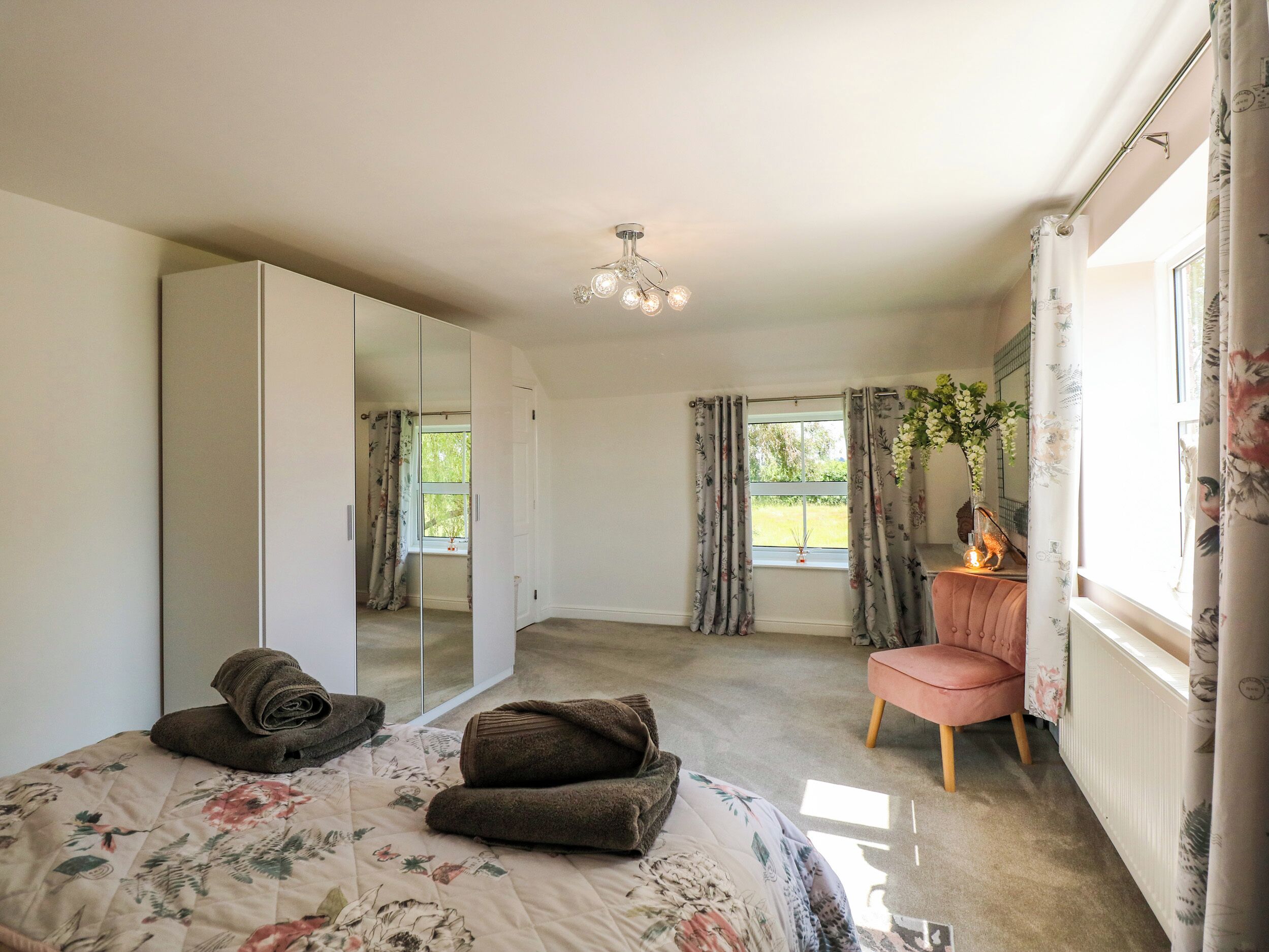 A bedroom with a bed, a mirror, a pink chair, and a window with curtains.