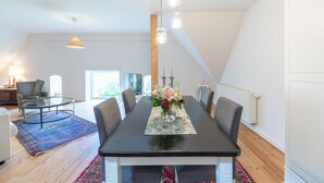 Uma sala de jantar com uma mesa comprida, cadeiras e luminárias pendentes. Há uma janela que oferece luz natural e vista para o verde lá fora.