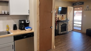 Een keuken met een vaatwasser, koffiezetapparaat en een schaal met chips. Er is een houten deur en een open haard met een televisie erboven.