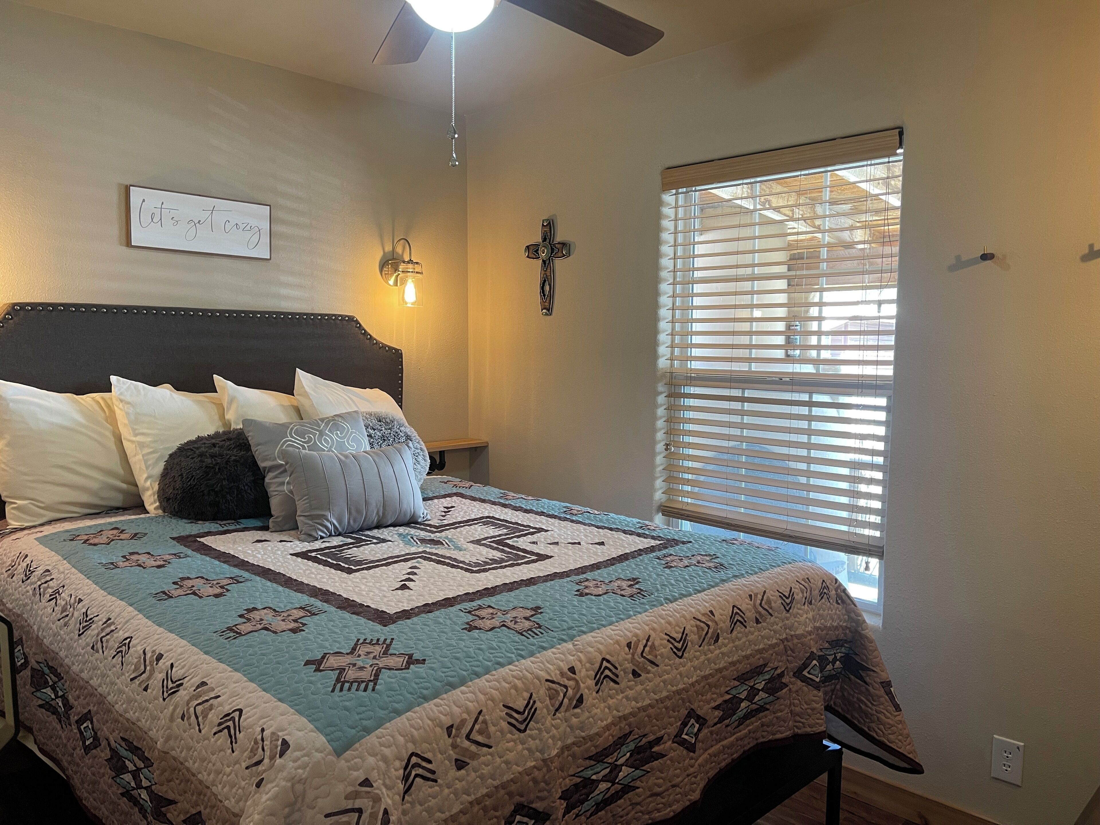 A bedroom with a bed, a ceiling fan, a wall-mounted light, a window with blinds, and a wall decoration.