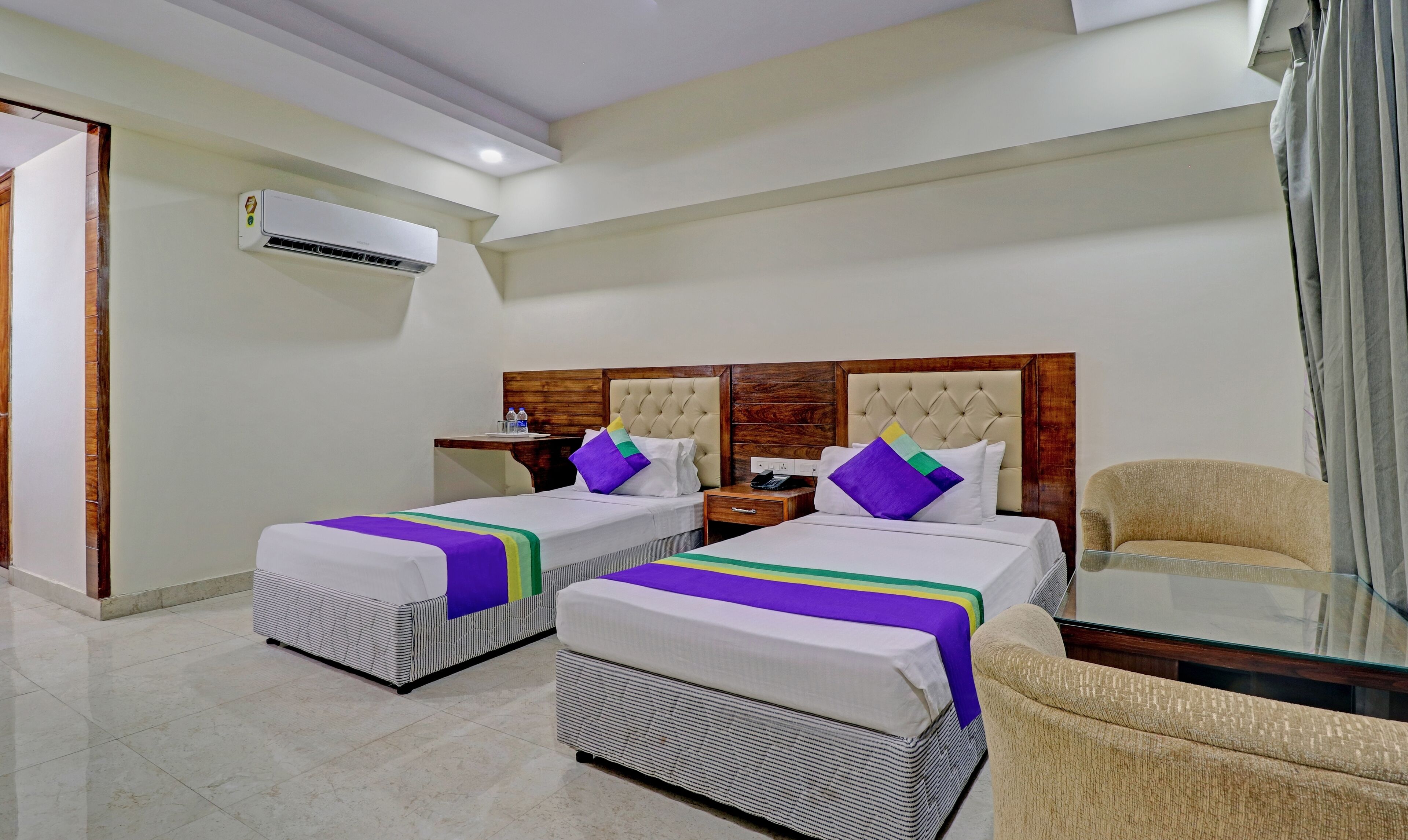 Habitación de hotel con dos camas, un sofá, una mesa de vidrio y un equipo de aire acondicionado.