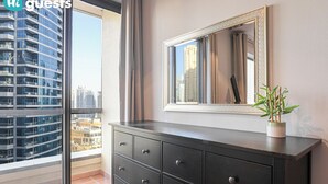A modern hotel room with a large window, a dark wooden dresser, a mirror, and a small potted plant.