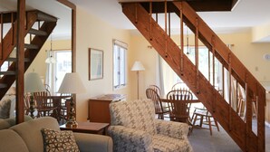 A living room with a wooden staircase, a sofa, patterned armchairs, a table, and a lamp.