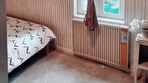 Een kamer met een schuine balkenlaag, een bed met een geborduurde spreideken, een raam met gordijnen, een radiator en een tafeltje met een bloempot.