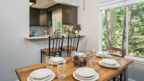 A dining area with a wooden table set for four, surrounded by chairs, a window with a forest view, and a kitchen area with cabinets and a window above the sink. - Home theater, hot tub, pool table, secluded, spacious. (Gatlinburg)