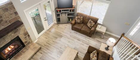 A spacious living room with a stone fireplace, a ceiling fan, a TV on a wooden stand, and a brown leather sofa with patterned cushions.