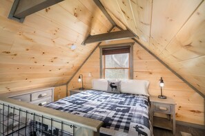 Ein gemütliches Schlafzimmer im Dachgeschoss mit schrägverkleidetem Holzboden, einem Bett mit kariertem Quilz, zwei Nachttischlampen und einem Fenster mit Blick auf Bäume.