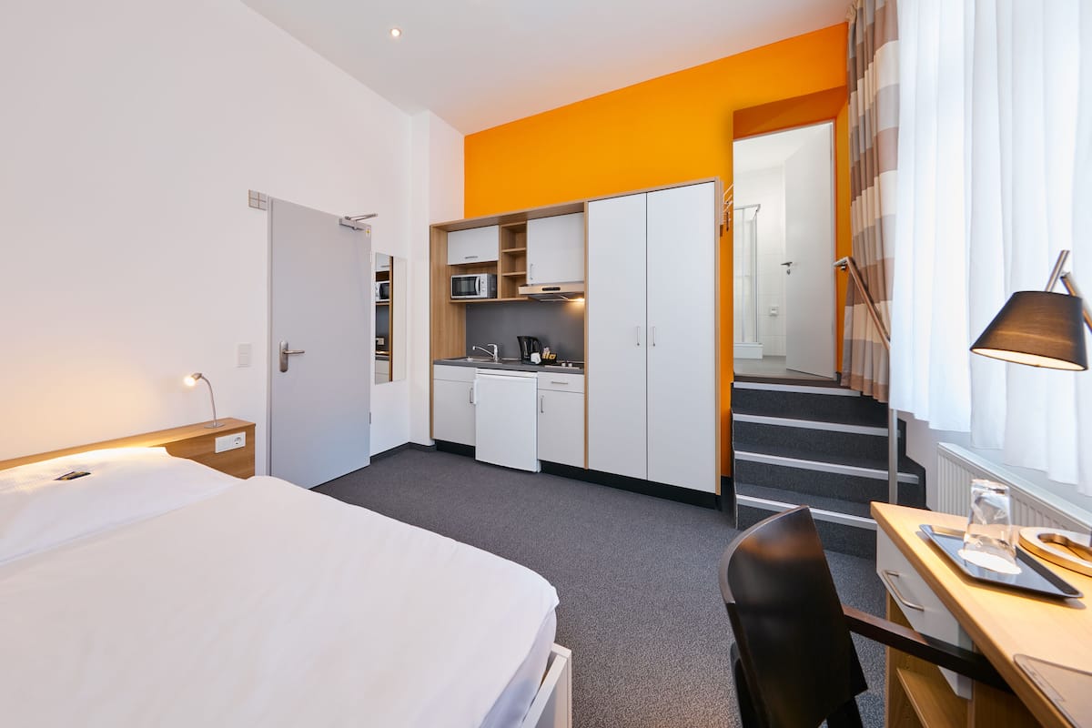 A hotel room with a bed, a desk, a chair, a kitchenette, and a staircase leading to another room.