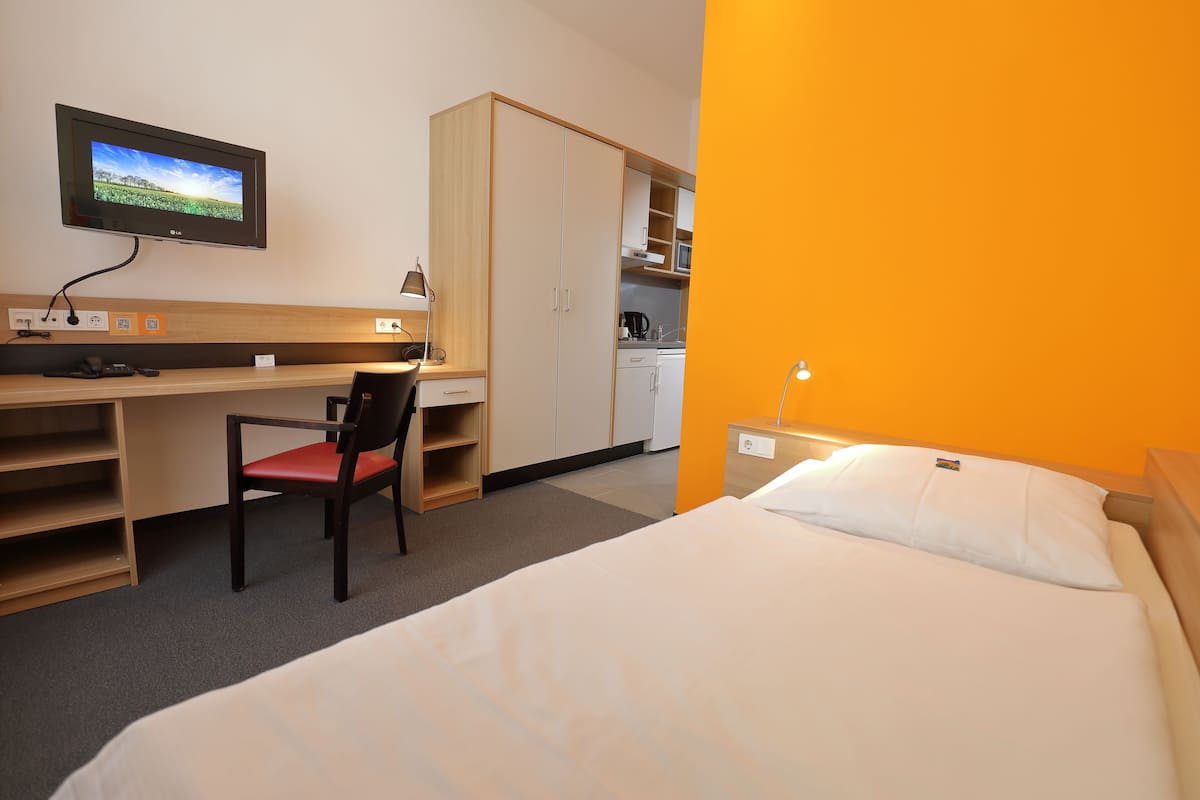 A hotel room with a bed, a desk with a chair, a television, and a kitchenette.