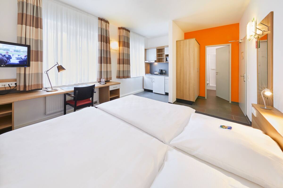 A hotel room with a bed, desk, chair, TV, and kitchenette.