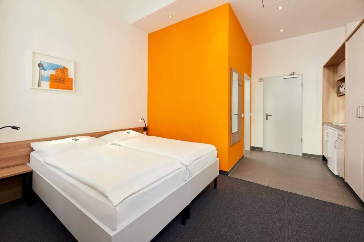 A modern hotel room with a double bed, a small desk, a chair, a bedside table, a mirror, and a view of the adjacent kitchen.