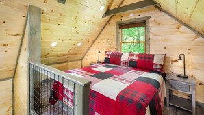 Een gezellig slaapkamer op zolder met een houten plafond, een groot bed met een quilt in rood en zwart, een raam met uitzicht op bomen en een klein nachtkastje met een lamp.