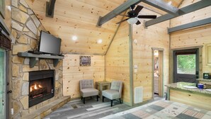 Interior acollidor d'una cabana amb una llar de pedra, una televisió penjada a una paret de pedra, dues cadires, una taula petita i una zona de cuina amb taulell i rentapelats.