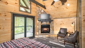 Una habitación de cabaña acogedora con chimenea de piedra, un televisor montado encima, cama con ropa de cama escocesa, dos sillones de cuero y un ventilador de techo.