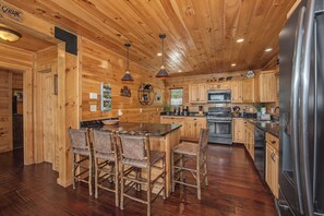 A cabin-style kitchen with wooden walls, a central island with bar stools, stainless steel appliances, and a hanging light fixture.