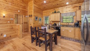Interior de una cabaña de madera con una mesa de comedor, sillas, una zona de cocina y un refrigerador.