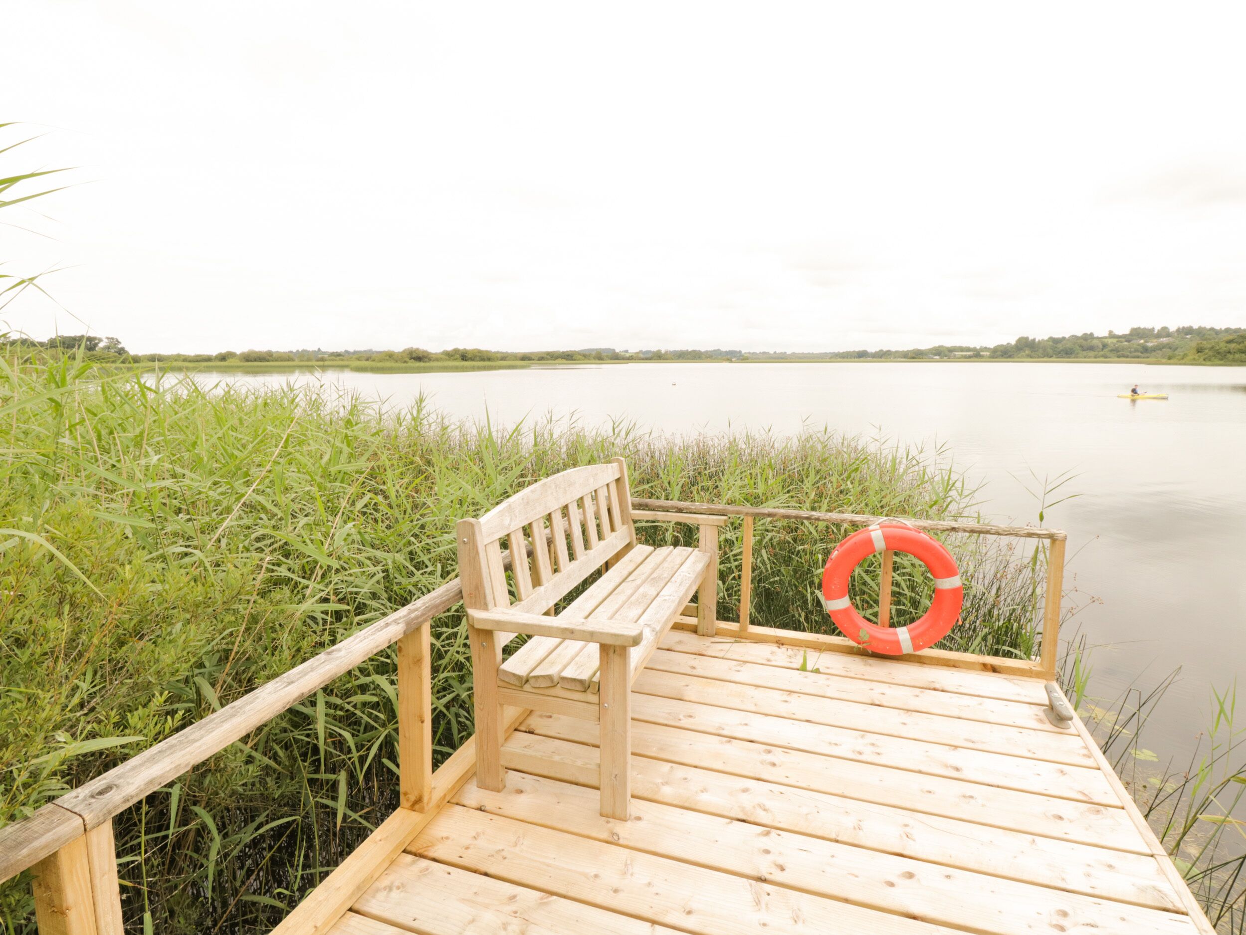 Terraza de madera con un banco y un flotador salvavidas, con vista a un lago tranquilo con carrizos y una persona remando a lo lejos.