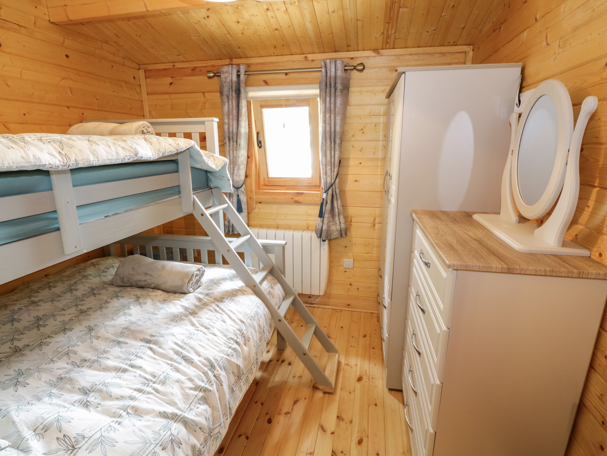A wooden cabin room with bunk beds, a dresser, a mirror, and a window with curtains.