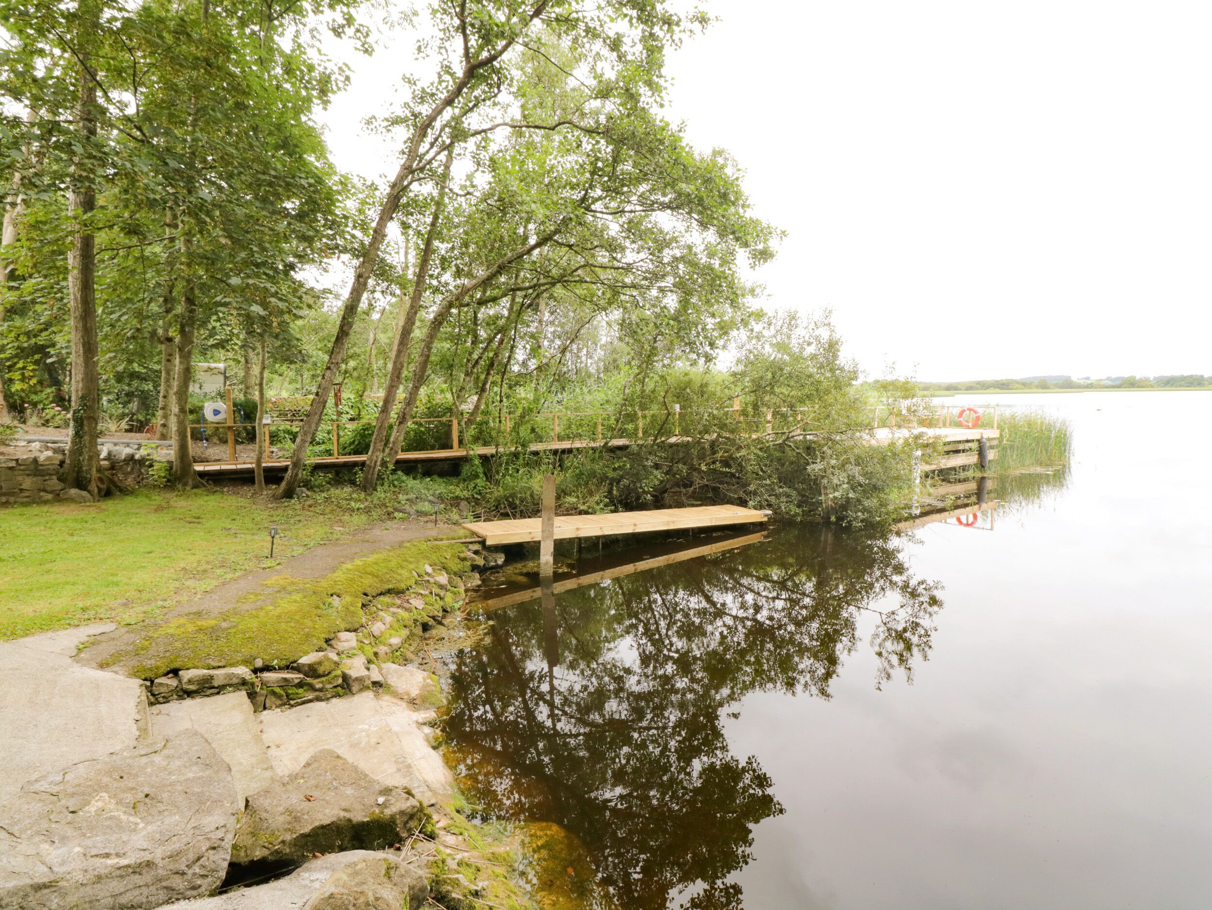 A calm lake with a wooden dock extending into the water, surrounded by lush greenery and trees.