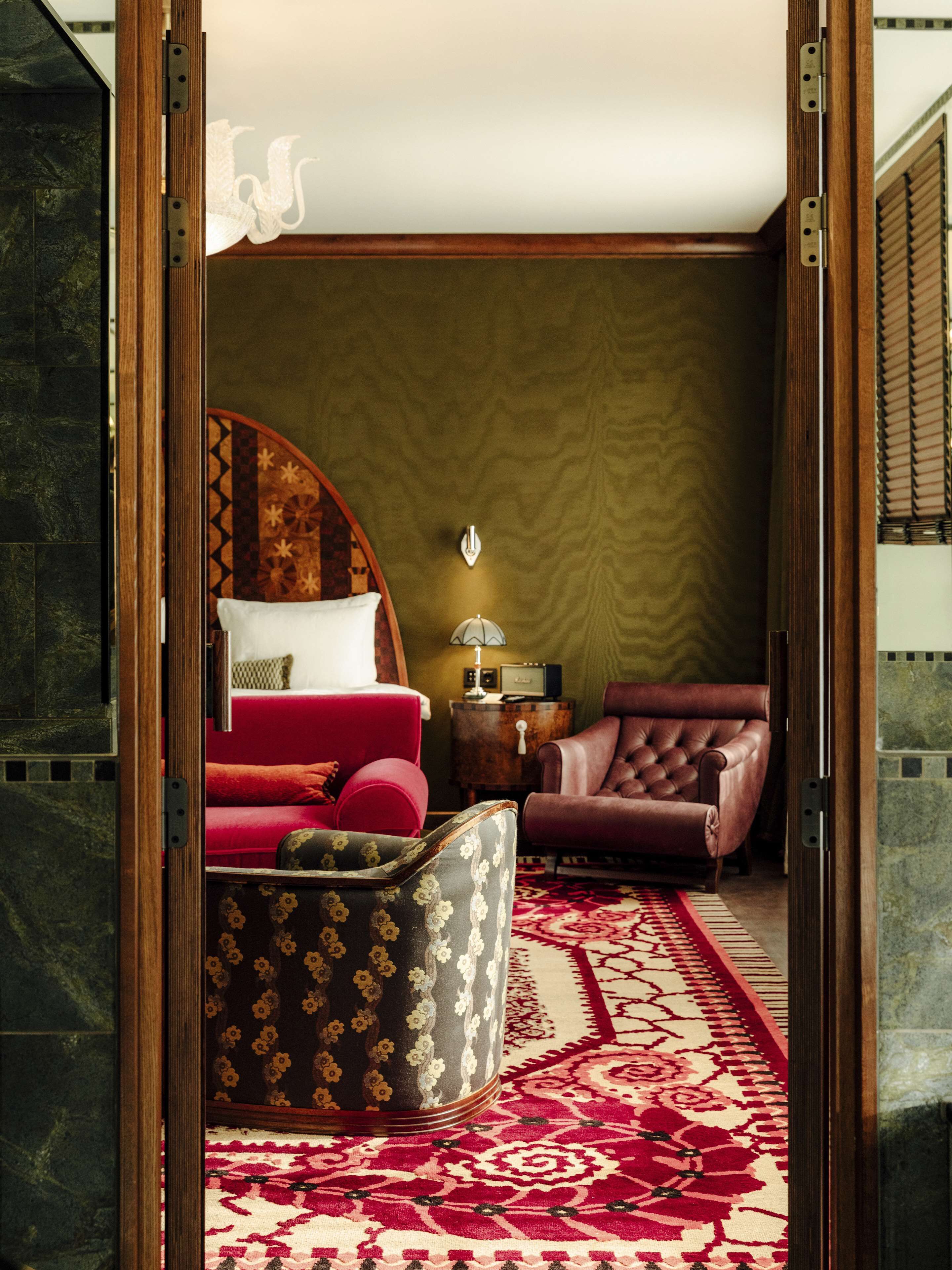 A room with a bed, a red sofa, a brown leather armchair, a patterned ottoman, a small table with a lamp, and a patterned rug.
