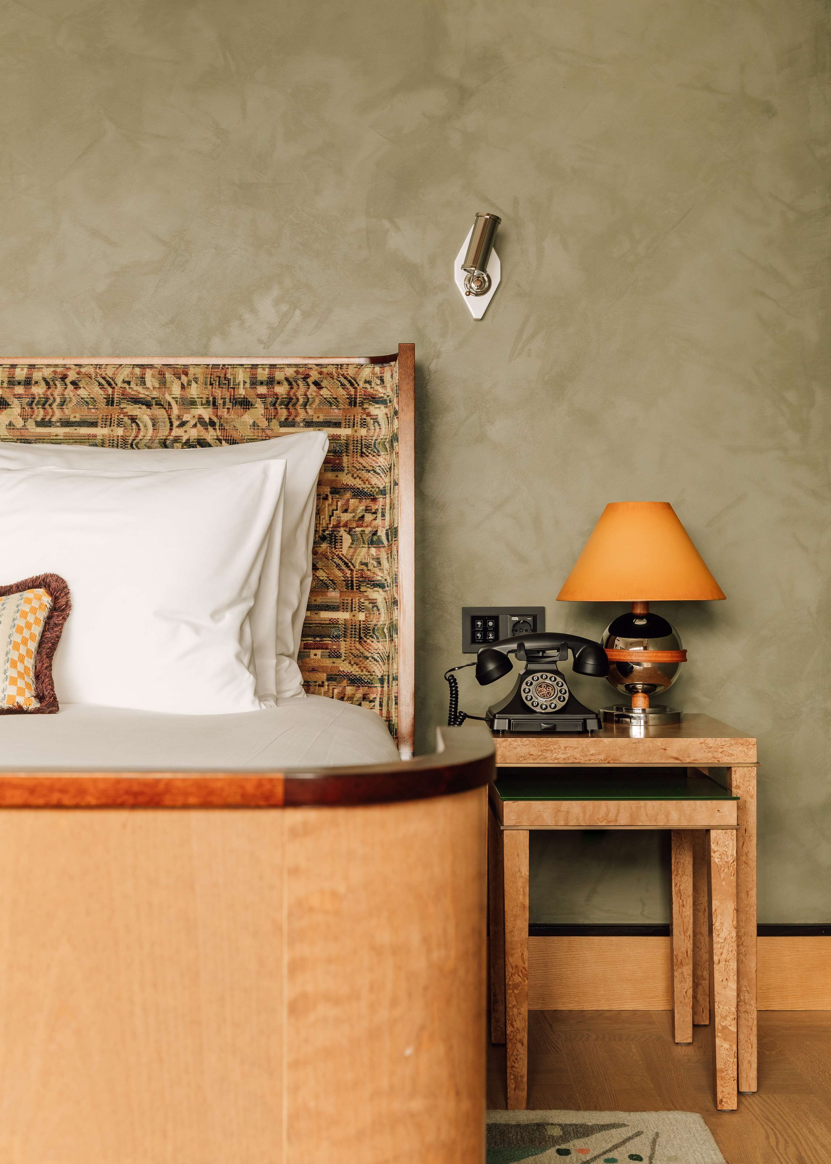 A bed with a patterned headboard, a bedside lamp, a telephone, and a wall-mounted light fixture.