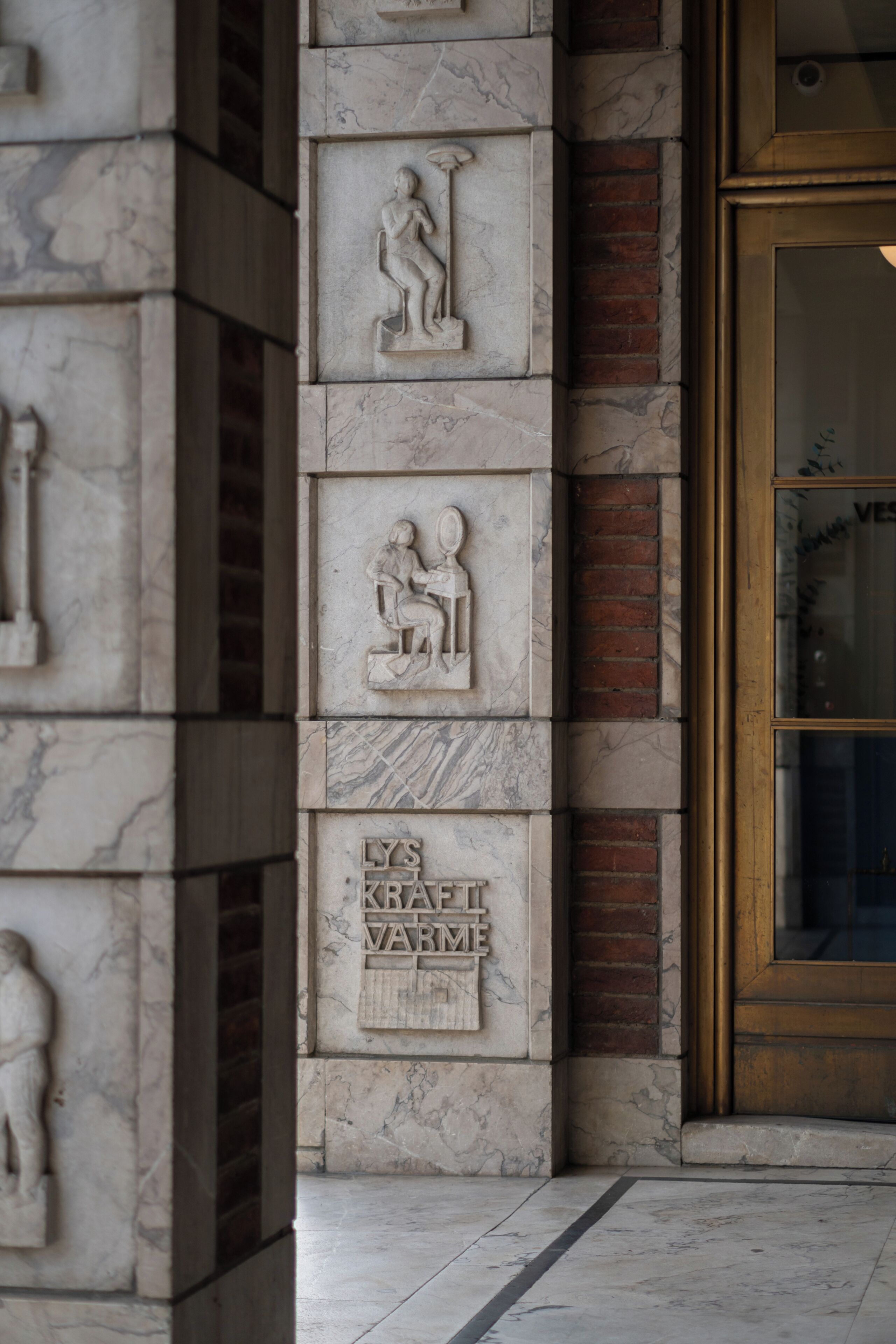 A building facade with marble relief sculptures, a brick column, and a glass door with a golden frame.