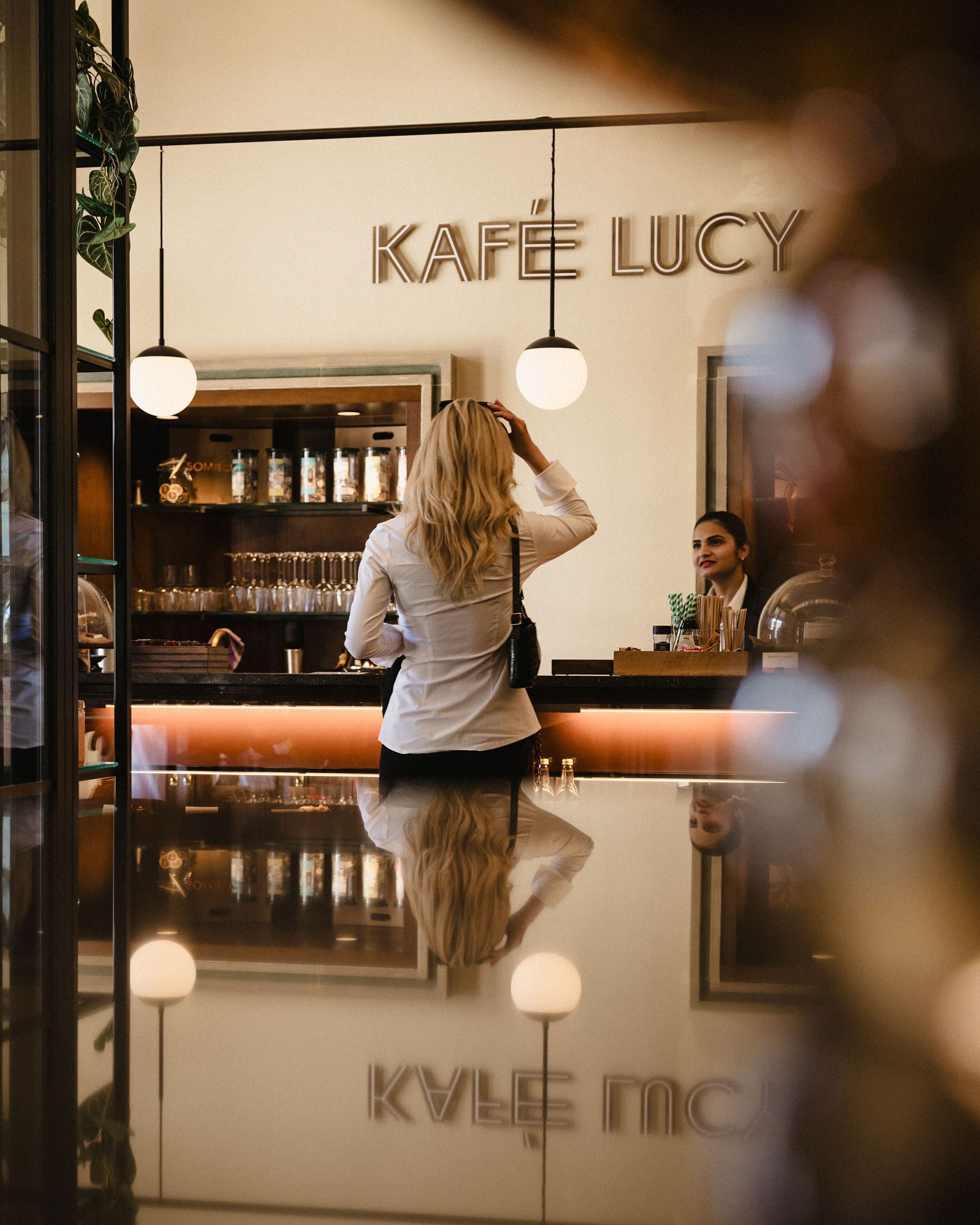Una mujer está de pie junto a una barra en una cafetería llamada 'KAFÉ LUCY'.