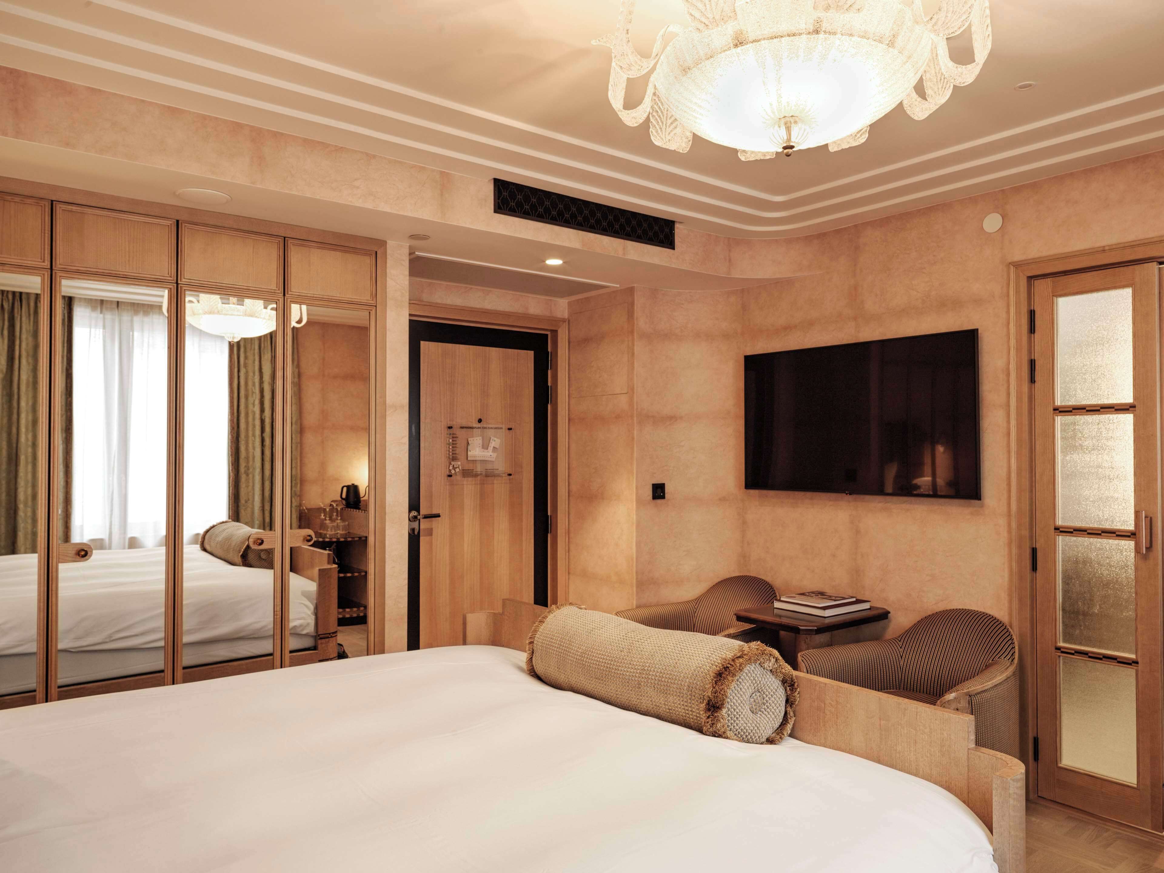 A hotel room with a large bed, a television mounted on the wall, a seating area with a sofa and chairs, and a wooden wardrobe with a mirror.