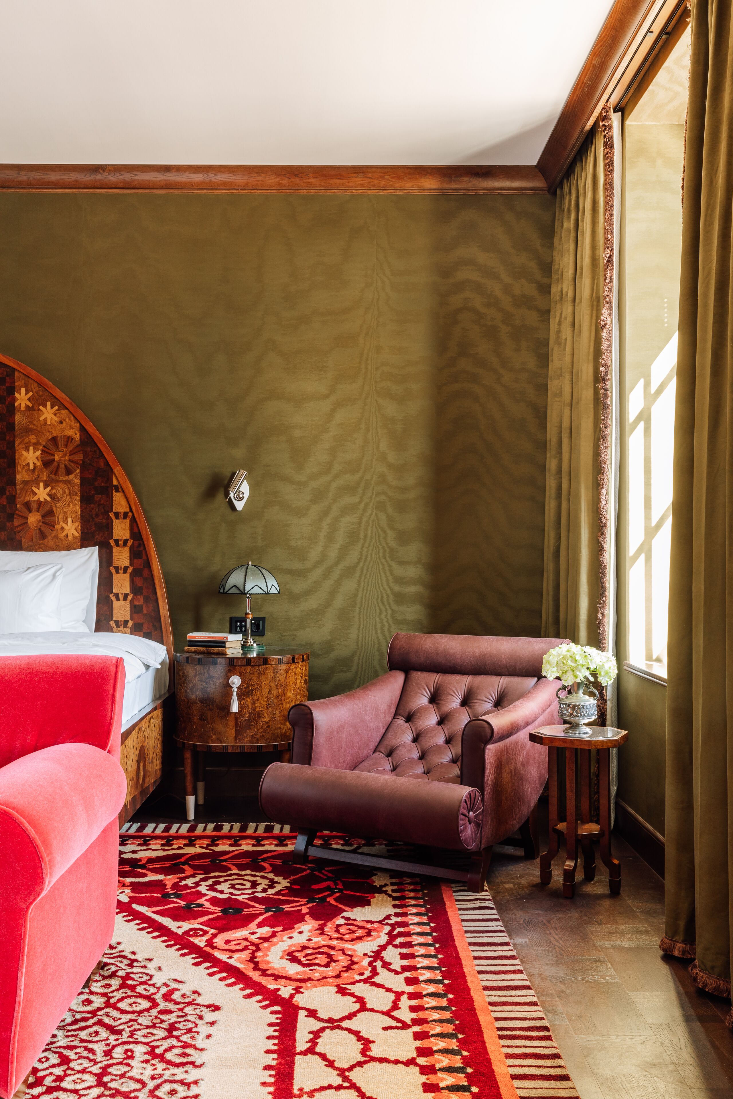 Una habitación con una cama, un sillón rojo, un sillón de cuero marrón, una mesita de noche con una lámpara y una ventana con cortinas.