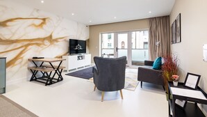 Un salon moderne avec un mur en marbre, un téléviseur à écran plat, un fauteuil gris, un canapé et un coin repas avec une table et un banc en bois.