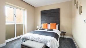 Une chambre à coucher moderne avec un grand lit, un banc, une petite table et une fenêtre donnant sur un bâtiment en briques.