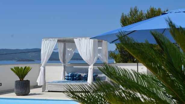 Una cama con cortinas blancas y almohadas azules, un paraguas azul y vistas al mar.