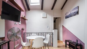 Uma sala de jantar com uma mesa redonda, cadeiras brancas, um ventilador e uma TV na parede.